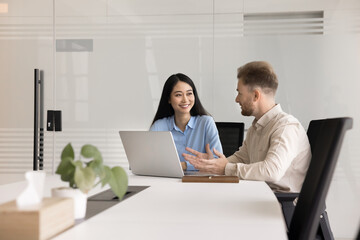 Couple of happy diverse business colleagues discussing online startup at laptop, talking, smiling. Project managers sitting at large table in office meeting room, using computer for teamwork,