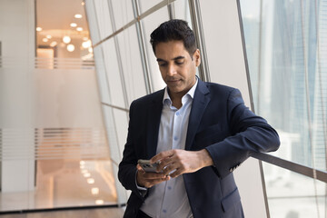 Focused middle aged Indian businessman in formal clothes using online bank application for financial transactions, typing on smartphone for work communication, standing at office window indoors