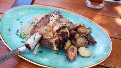 Overhead view of roasted pork knuckle with crispy skin and roasted potatoes on a blue plate at an outdoor restaurant, with beer in the background