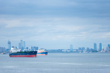 Obraz premium A cargo ship rests anchored in the bay, dominating the foreground. In the distance, the steel-blue skyline of New York City looms under a cloudy sky, blending into the moody, overcast atmosphere.