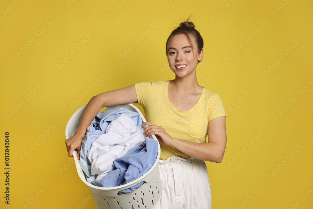 Wall mural Happy young housewife with basket full of laundry on yellow background