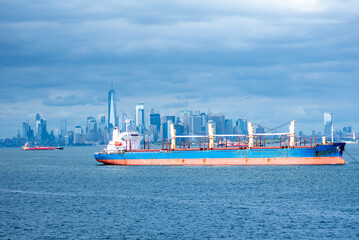 Obraz premium A cargo ship rests anchored in the bay, dominating the foreground. In the distance, the steel-blue skyline of New York City looms under a cloudy sky, blending into the moody, overcast atmosphere.