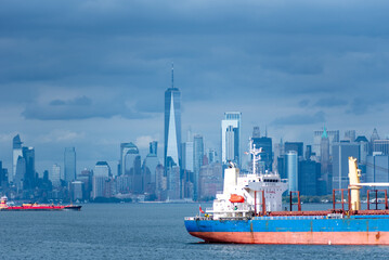 The ship's stern takes focus as it glides through the bay, with New York City's skyline blurred in the background. The cloudy sky casts a soft, steel-blue tone over the entire scene.