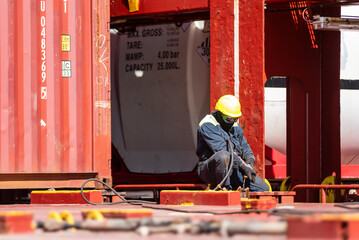 A crew member kneels on the deck of a merchant ship, using a pneumatic tool to remove rust. Focused...