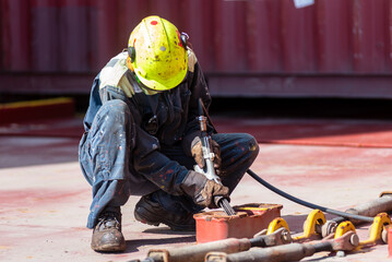 A crew member kneels on the deck of a merchant ship, using a pneumatic tool to remove rust. Focused...
