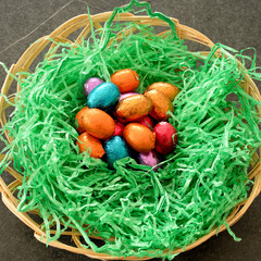 Close up on Easter Nest standing on black table, with colorful shiny chocolate easter eggs, and green grass imitation.