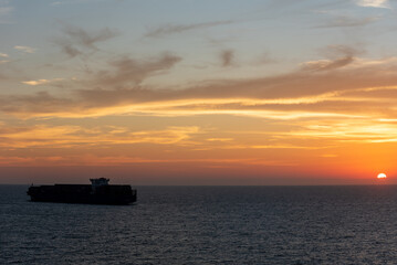 The calm silhouette of a massive container ship gliding across the smooth sea, enveloped by the golden hues of the sky as the sun bids farewell to the day.