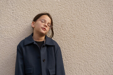 Little sad girl with ponytail and glasses stands facing the camera against the wall. Smart and shy...