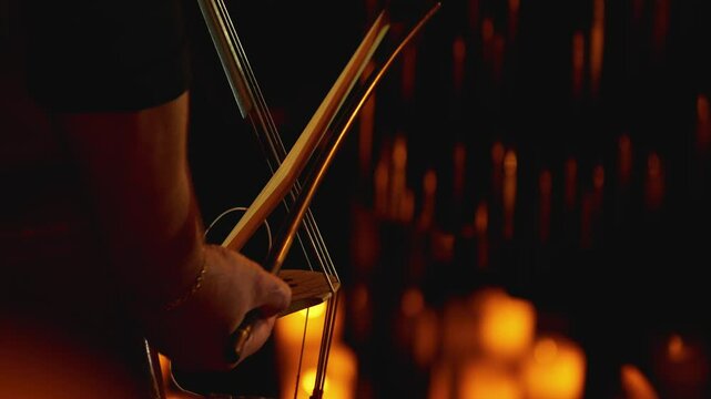 A close-up of a musician's hand using a bow to strike the strings of a double bass by candlelight. The hues of the instrument's wood contrast with the warm glow of the flame. A candlelight concert