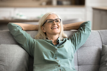Satisfied older woman wear glasses casual blouse resting leaned on soft cozy couch in living room,...