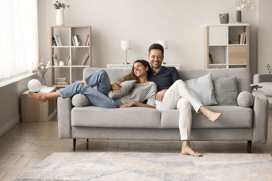 Positive happy young husband and wife relaxing on comfortable couch together, talking, hugging, laughing, having fun, enjoying romantic leisure, relaxation at home, good warm relationships
