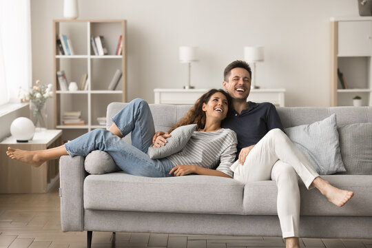Positive happy young husband and wife relaxing on new comfortable pale grey sofa, hugging, talking, laughing, having fun, spending home funny leisure time together