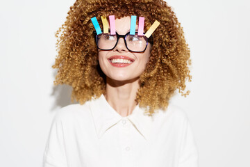 Cheerful woman with curly hair and colorful clips on glasses, wearing a white shirt, against a...