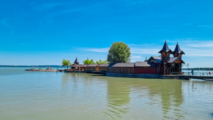 Keszthely Island Bath on Lake Balaton, Hungary. Lakeside scene with unique wooden structure. Multiple towers with pointed roofs and ornate details. Unique bathhouse or spa complex. Walkway from shore