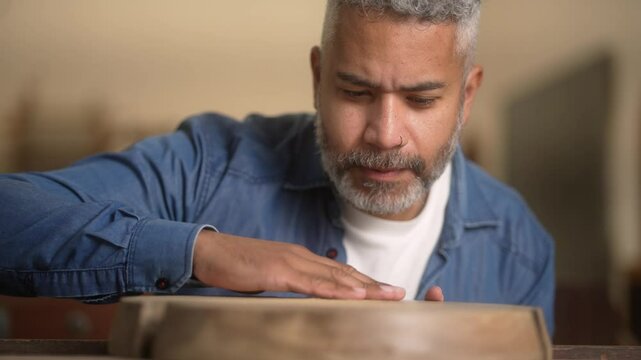 Focused craftsman carefully inspecting wooden piece in workshop. Skilled artisan blowing dust off woodworking project with attention to detail and pride