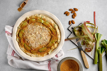 gluten free swiss rhubarb tart, rhabarberwahe, sprinkled with chopped almonds and icing sugar in baking dish nd ready for oven on gray table with ingredients
