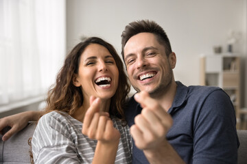 Cheerful happy couple looking at camera, laughing, showing finger heart shapes, symbol of love, happiness, romance. Husband and wife relaxing on home couch together, having fun