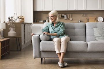 Obraz premium Elderly 60s woman on retirement using laptop at home, sit comfortably on sofa, browsing wireless internet, communicates with family or friends through email or social media, shopping or working online