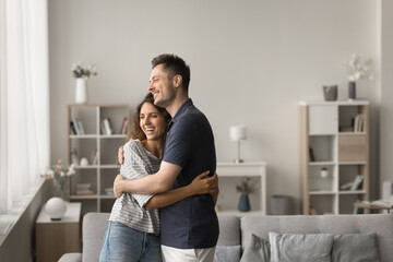 Happy young family couple of homeowners standing in comfortable home interior, hugging in new...