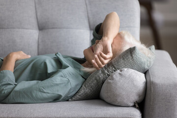 Older woman lying on sofa cushion, feeling fatigued, unmotivated, taking rest at home. Peaceful aged female having midday break, daytime nap indoors, relieving stress and mental or physical exhaustion