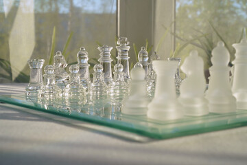 Glass chess on a white table in a bright, sunlit room. Space for text. The concept of hobby and leisure. Eye level view.