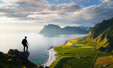 Hiker with backpack stands at the edge of rocky mountain fjord,overlooking a sunny valley below.Tourist standing on top of mountain, enjoying the peak of high rocks and an active lifestyle in Lofoten