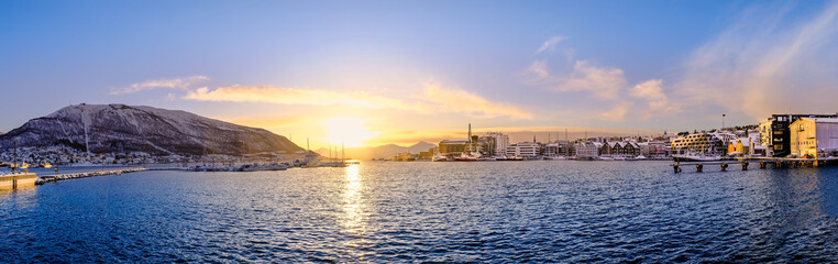 Fototapeta premium Panorama of Tromso harbor at sunset in winter, Norway. Scandinavian buildings and boats in port of Tromso and Storsteinen mountain. Snowy winter landscape in arctic circle town in far northern Norway