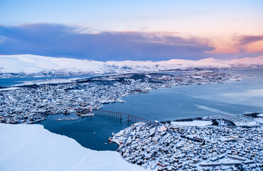 Aerial view of Tromso, Norway at sunset in winter from Storsteinen mountain. Tromsoya island with snowy roofs of Tromso downtown, harbor, Sandnessundet Bridge over Fjord and mountains in background