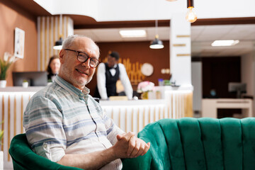 Portrait shot of senior man seated on cozy sofa in exclusive hotel reception, waiting for check-in procedure. Comfortable lobby offers relaxing lounge area for elderly male traveler looking at camera.