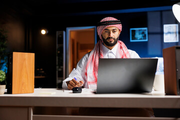 Middle Eastern man wearing Arabian clothing is seated at the table, engrossed with research on his...