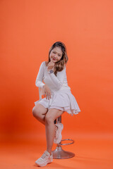 full body teenage girl white sweater cute smile slanted right hand hold chin chair sitting on hydraulic chair, standing on orange background. for fashion, lifestyle, beauty concept