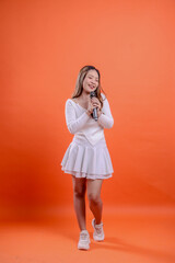 full body beautiful indonesia woman white top standing singing cheerfully close eyes both hands holding mic isolated orange background. for fashion concept, advertising, lifestyle, entertainment