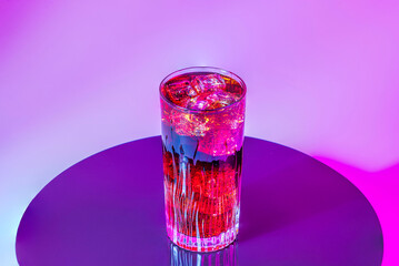 Tall glass of cold beverage with ice cubes against vibrant pink and purple light on black reflective surface.