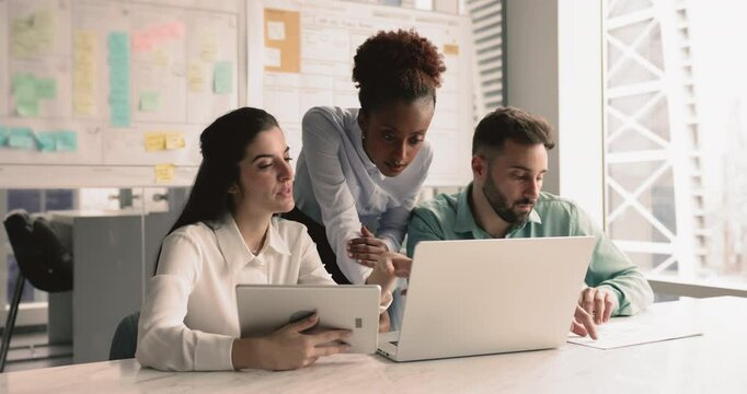 Three focused workmates, board of directors met in office for briefing event, discuss report using laptop and tablet device, check data in papers, share opinion, learn marketing campaign efficiency