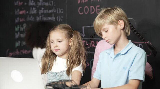 Smart girl using laptop programing engineering code with diverse friends ay table with microchip and main board scatter around in STEM classroom at blackboard written with coding prompt. Erudition.