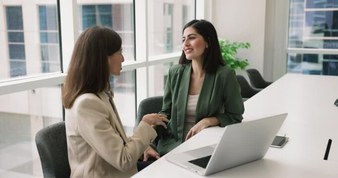 Two multiethnic young businesswomen meet in conference room, discussing strategic plans, commercial offer, making deal, shaking hands to finalize their agreement and partnership. Business, negotiation