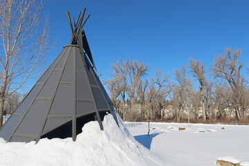 Modern metal and glass tipi teepee tepee in the snow with leafless cottonwood trees and a blue sky background