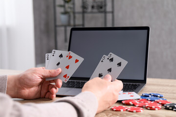 Online poker. Man with playing cards, chips and laptop at wooden table indoors, closeup