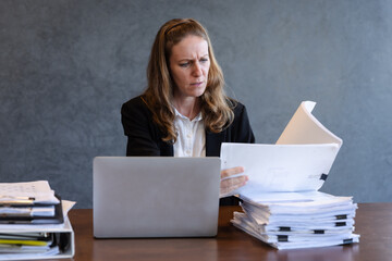 Focused Businesswoman Reviewing Documents in Modern Office Workspace, Surrounded by Paperwork and Using Laptop for Project Management