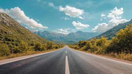Naklejka premium A straight asphalt road lined with green trees and mountains, under a bright sky with scattered, fluffy white clouds.