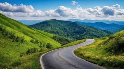 Fototapeta premium A winding asphalt road through green mountain valleys, with a clear blue sky and soft, fluffy clouds on a sunny day.