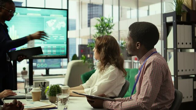 Diverse group of professionals meeting to discuss a new development strategy, utilizing infographics and monthly analysis numbers to reach target and accomplish organizational goals. Camera B.