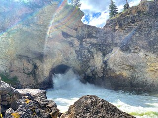 Waterfall coming out of the side of a rocky cliff with pine trees and river 