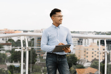 Confident City Planner: Young Asian businessman reviewing documents on tablet, standing on rooftop...