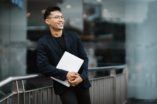 Confident and Connected: A young professional, radiating optimism, leans against a railing, laptop in hand, gazing towards a bright future. The urban backdrop underscores his ambition and drive.