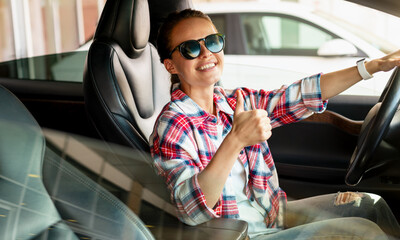 A young female sits in her new car at the dealership, smiling and giving a thumbs-up sign. Car...