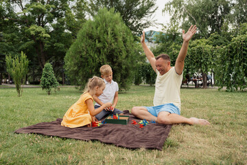 Father and children playing a board game during a picnic in a park. The family is enjoying quality time together outdoors.