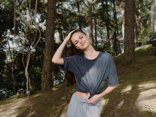 Cheerful young woman in casual clothing standing outdoors, touching her hair, surrounded by lush greenery Nature and carefree lifestyle concept