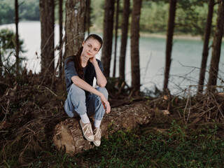 Naklejka premium Thoughtful young woman sitting on a log near a serene lake, dressed casually in gray attire, surrounded by lush greenery and tall trees Nature and relaxation concept