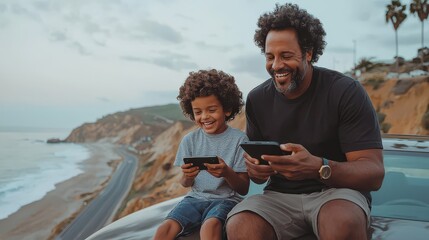 Father and son enjoying video games together on car hood with scenic coastal view, smiling and bonding in nature